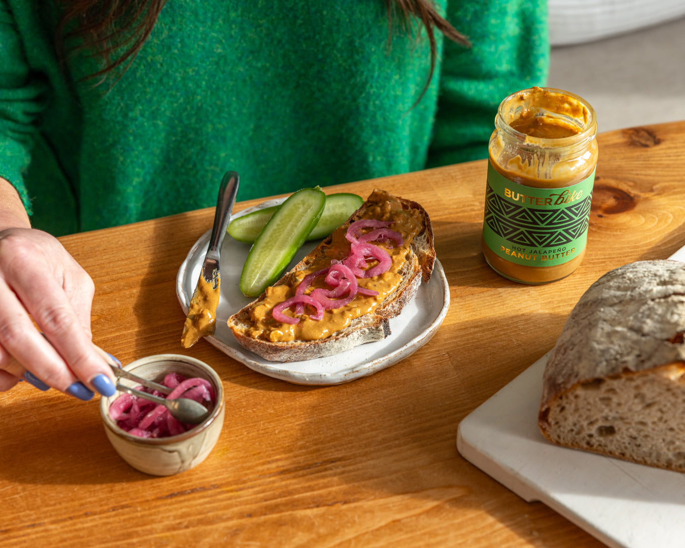 Person spreading peanut butter on bread with pickles and pink onions, set on a wooden table.